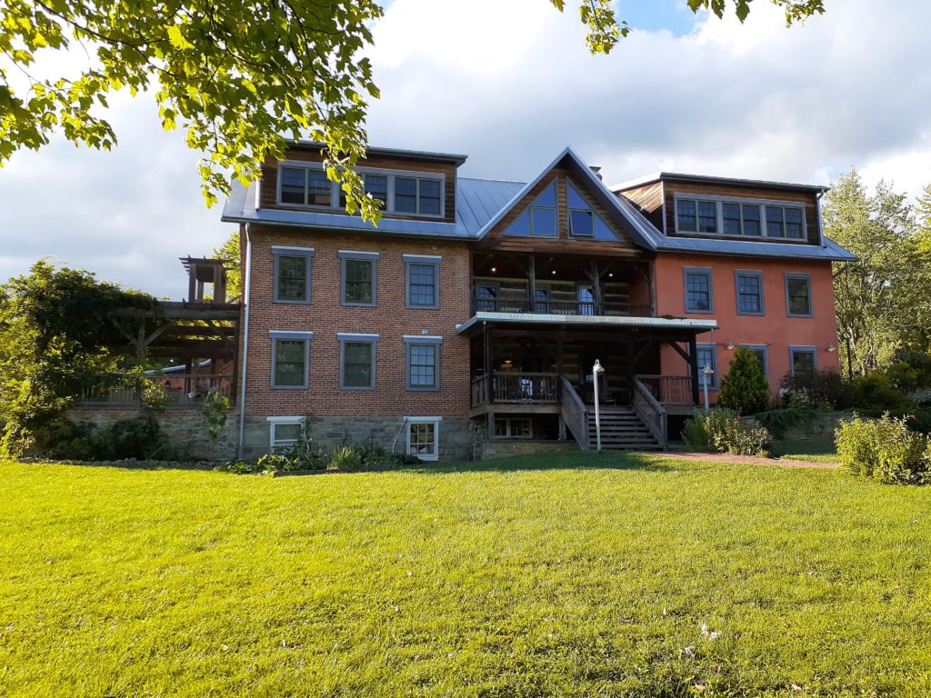 Writing with the support of a writing community. A picture of a multi-story red brick building, with a large porch covered in vines sits on a grassy lawn.